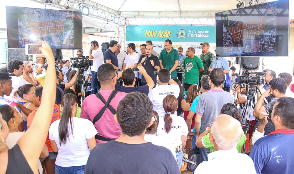 prefeito falando ao microfone em cima de palco com pessoas atrás e público assistindo em baixo à frente